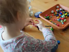 Dziecko z zapałem zajmuje się nawlekaniem koralików na sznurek, tworząc kolorową biżuterię.