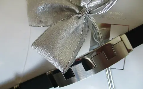 Elegancka bransoleta z czarnym paskiem i metalowymi elementami, ozdobiona srebrną kokardą. Idealnie pokazuje, jak zapakować biżuterię na prezent.