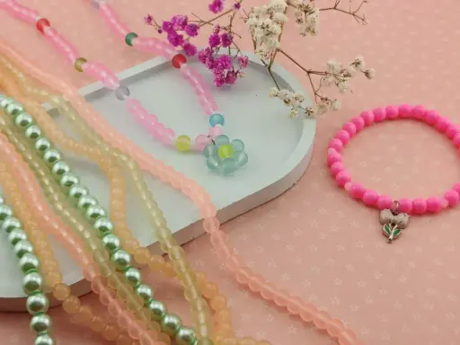 Kolorowe koraliki do bransoletek: naszyjnik z kwiatkiem, różowa bransoletka z zawieszką, zielone i pomarańczowe sznurki.